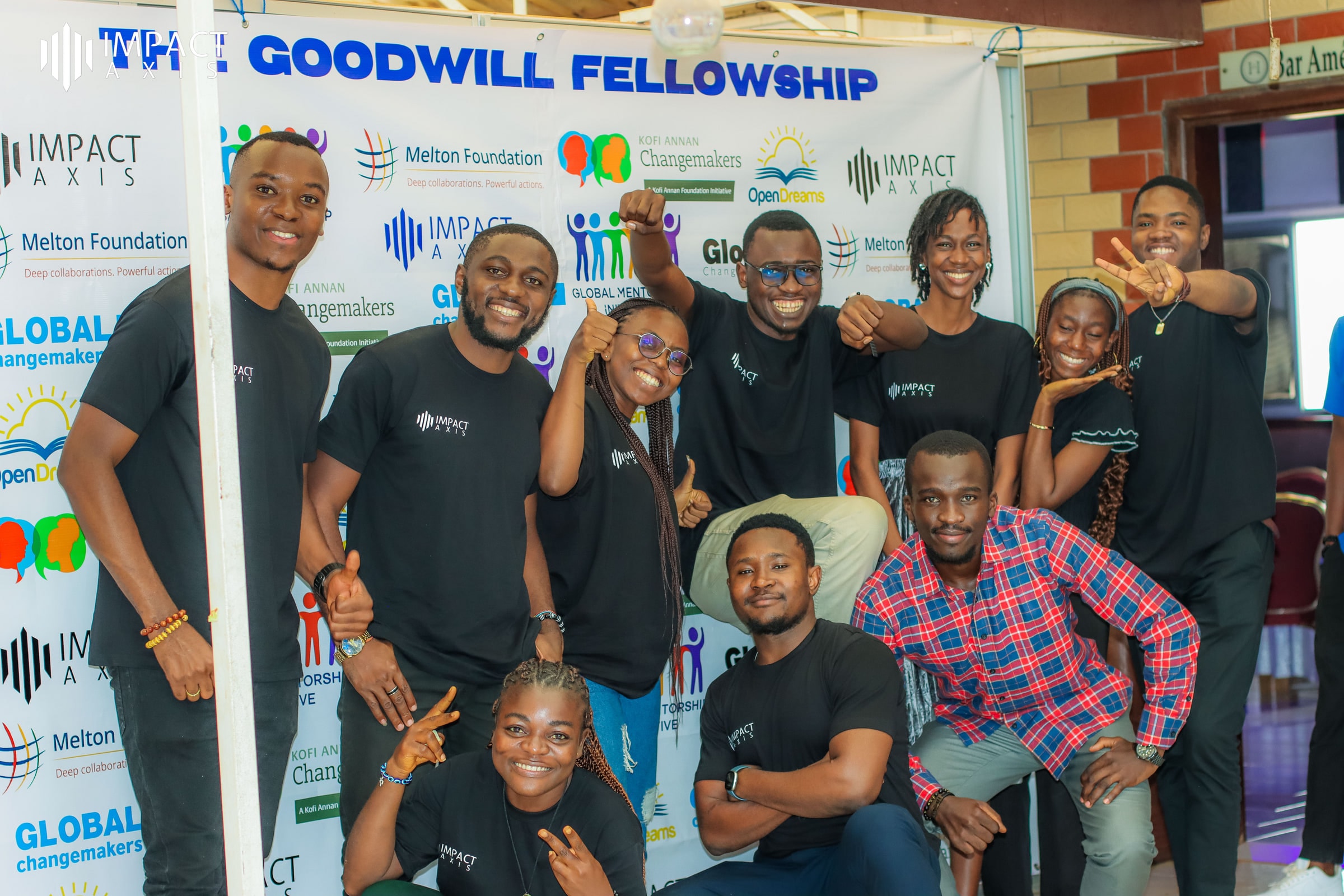 Goodwill Fellowship participants in black Impact Axis T-shirts posing together in front of the programme banner